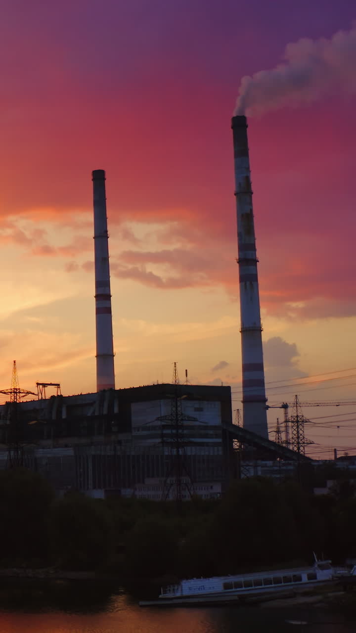 Industrial zone in the evening nature. Transmission lines, factory with pipe releases smoke near the river at sunset. View from the river. Vertical video