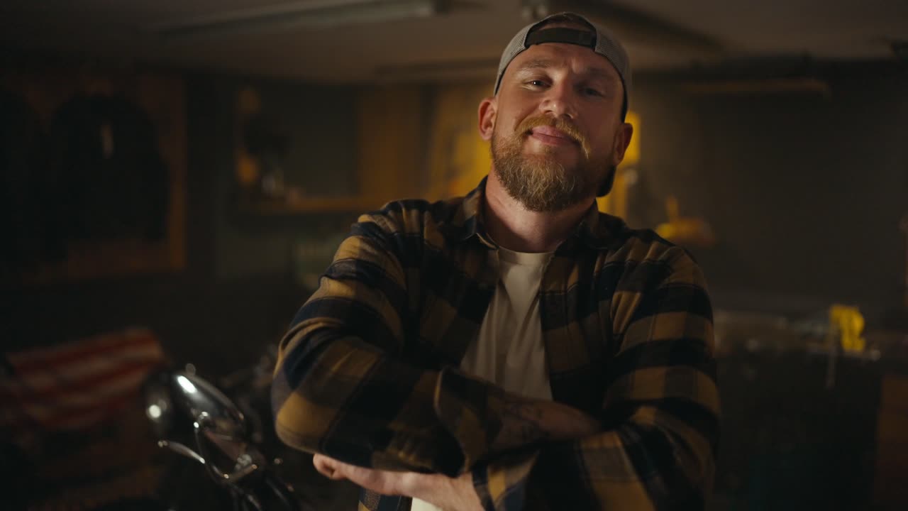 Portrait of blond biker with a beard who puts on a cap and poses in his motorcycle workshop