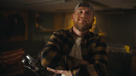 portrait of blond biker with a beard who puts on a cap and poses in his motorcycle workshop