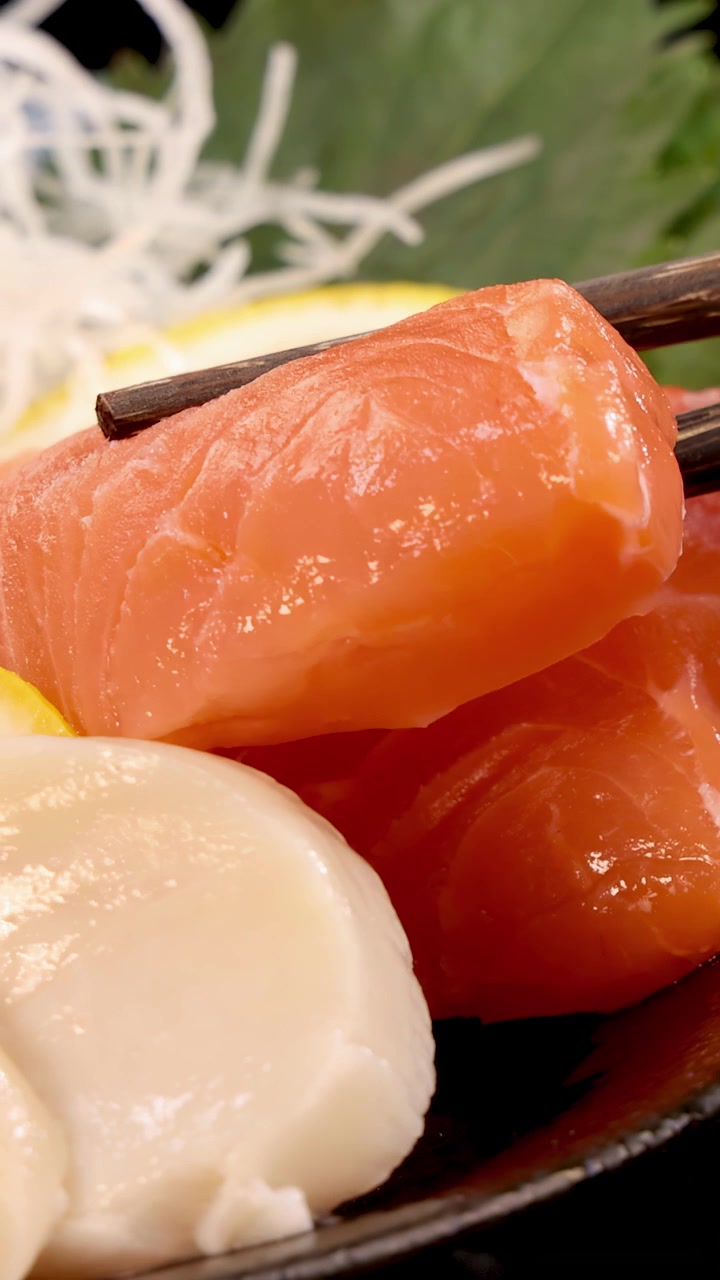Essstäbchen nehmen vorsichtig eine Scheibe frisches Lachssashimi von einem japanischen Sushi-Teller mit Jakobsmuschel, Zitrone und Shiso-Blatt unter heller Studiobeleuchtung auf