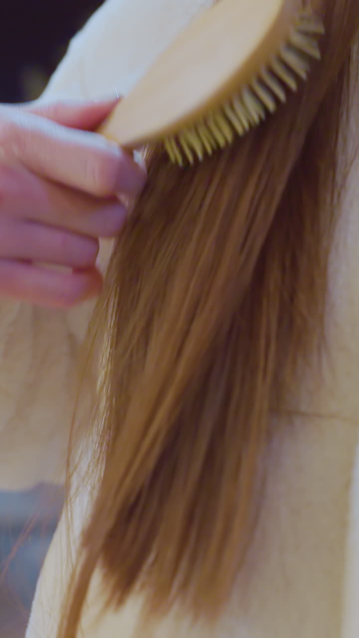 Close-up of woman in cozy white winter outfit brushing her long, straight hair with a wooden comb, soft, warm lighting enhances the scene, with a blurred background of an urban setting at night