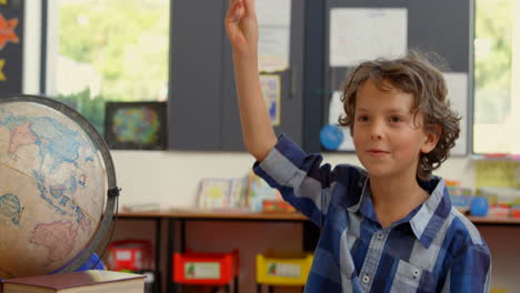 front view of caucasian schoolboy raising hand on desk in classroom at school 4k