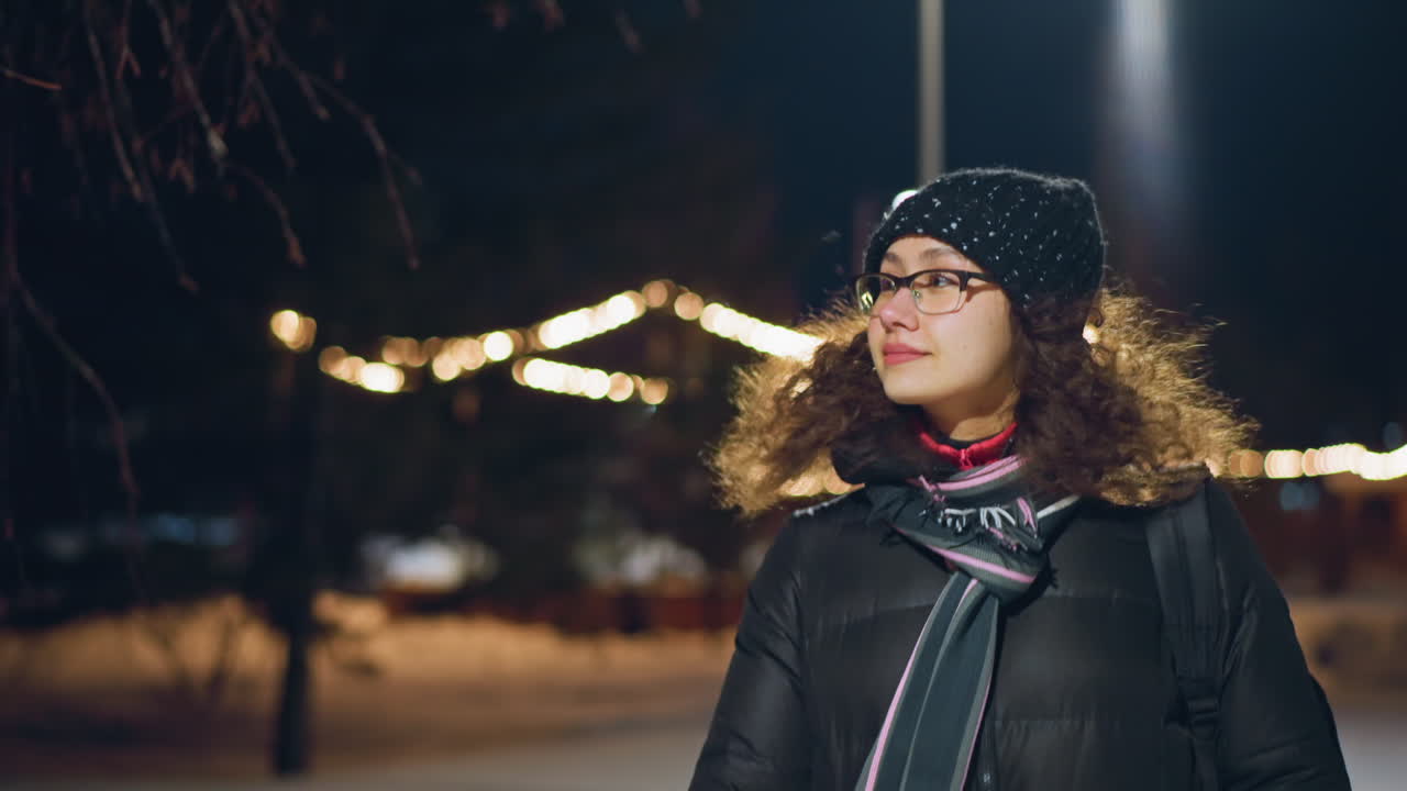 Woman wearing glasses winter coat scarf knitted hat walking outdoors at night illuminated by festive string lights looking thoughtful enjoying cold evening atmosphere with snow background and glowing bokeh