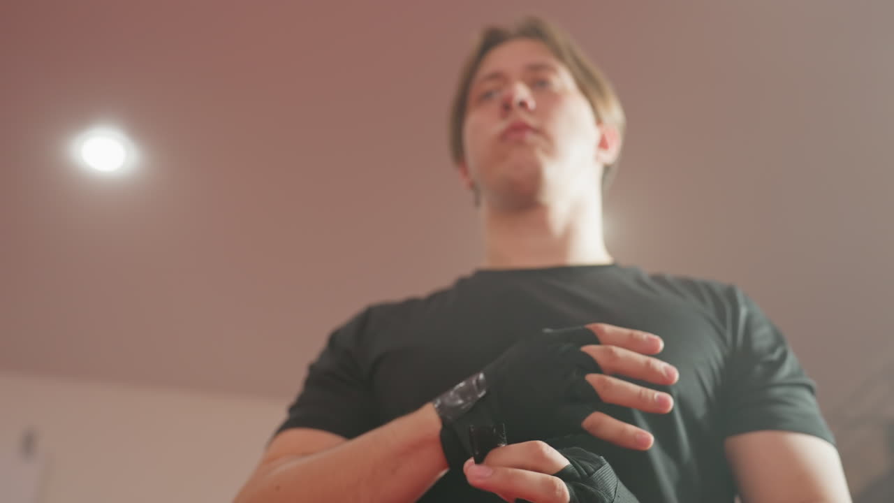 Athlete prepares for combat sport training by wrapping hands with protective black bandages, focusing on technique, discipline, and strength while wearing black sportswear inside brightly lit gym environment