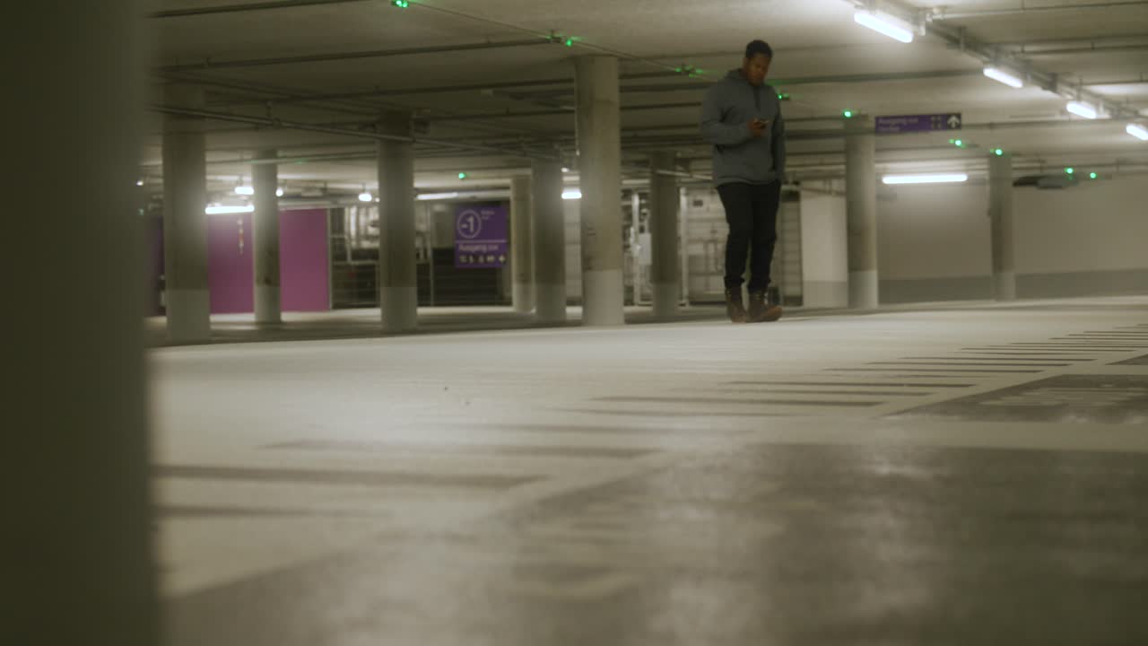 View of black male on phone walking inside empty underground parking lot with bright lights in Metzingen, Germany, Baden Wurttemberg, Europe, panning view angle