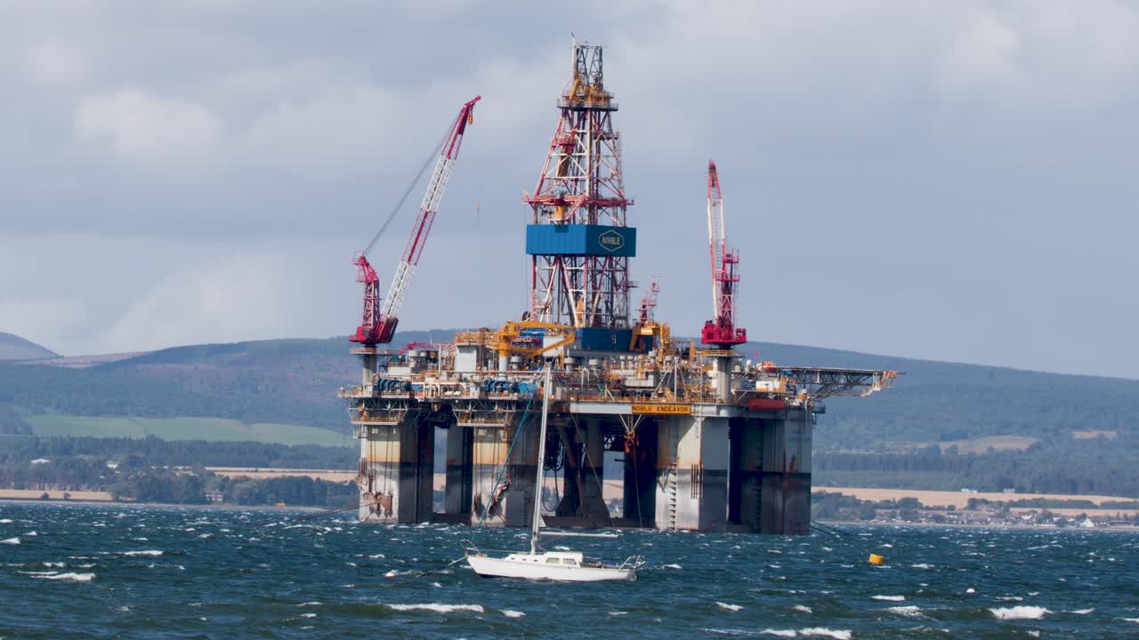 Hvid sejlbåd passerer stor offshore olieplatform på oprørt hav, dagslys, bredt billede.