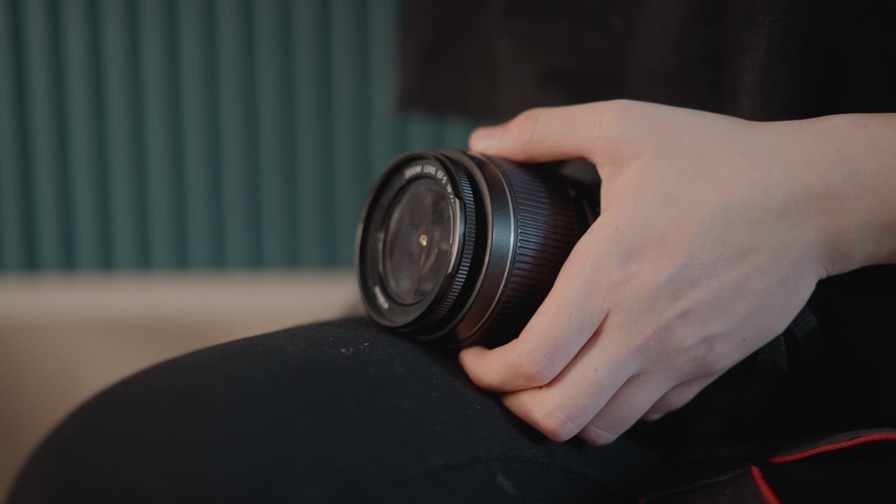 Close-up shows hands gripping camera lens while turning focus ring, creating visible twisting motion and careful inspection in calm indoor studio light with shallow depth and precise manual handling