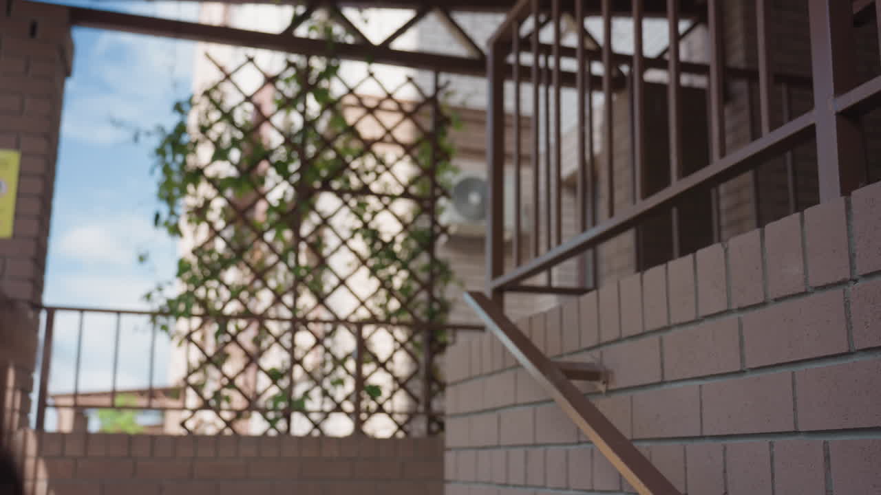 Una mujer de piel clara con un abrigo largo sube una escalera fuera de un edificio de ladrillo, vista trasera con bolso al hombro, barandillas de metal, celosía con hiedra verde, luz de día fría y movimiento ascendente decidido hacia el rellano