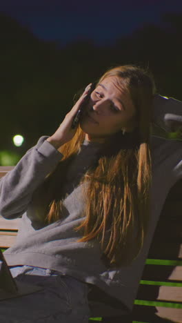mujer joven sentada en un banco del parque por la noche, ocupada en una llamada telefónica con la computadora portátil en el regazo, descansando la cabeza en la mano, la iluminación suave del parque destaca su expresión enfocada, rodeada de vegetación