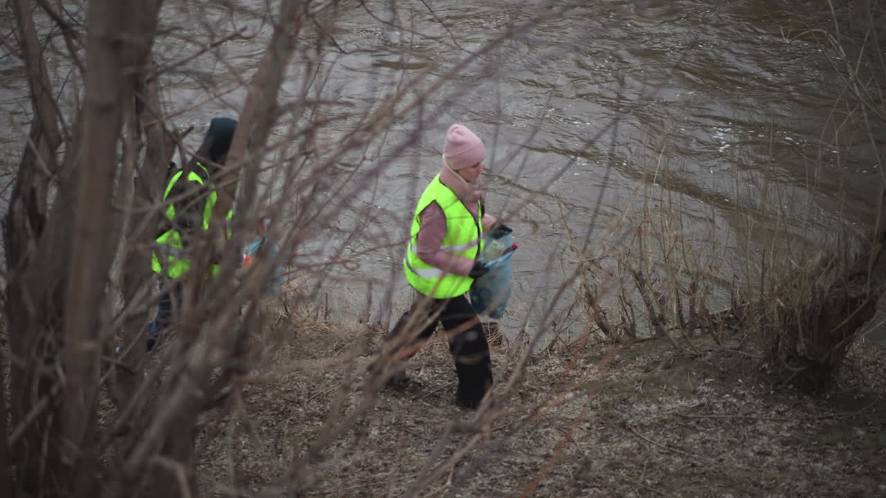 Volunteers in high visibility vests walking along riverbank collecting trash during environmental cleanup in early spring forest area with bare trees and cold weather conditions