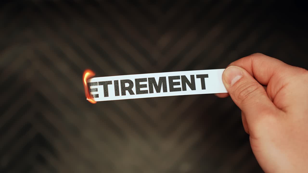 Close-up hand holds a paper strip reading “RETIREMENT” as the edge burns with a small flame, symbolizing financial risk, inflation, and uncertain future planning with copy space