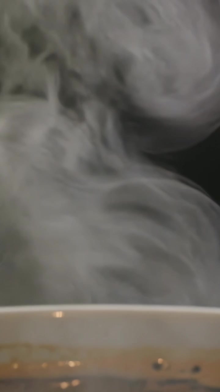 Vidéo verticale : Dégageant de la chaleur, une tasse à bord blanc avec une boisson foncée émettant une épaisse vapeur sur une table, avec des bulles.