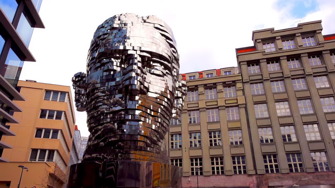 Prague, Czech Republic - November 2, 2021: Head of Franz Kafka stands proudly amidst the buildings. Clouds drift by in the blue sky, creating a striking contrast with the shiny metal. People walk nearby, admiring the art