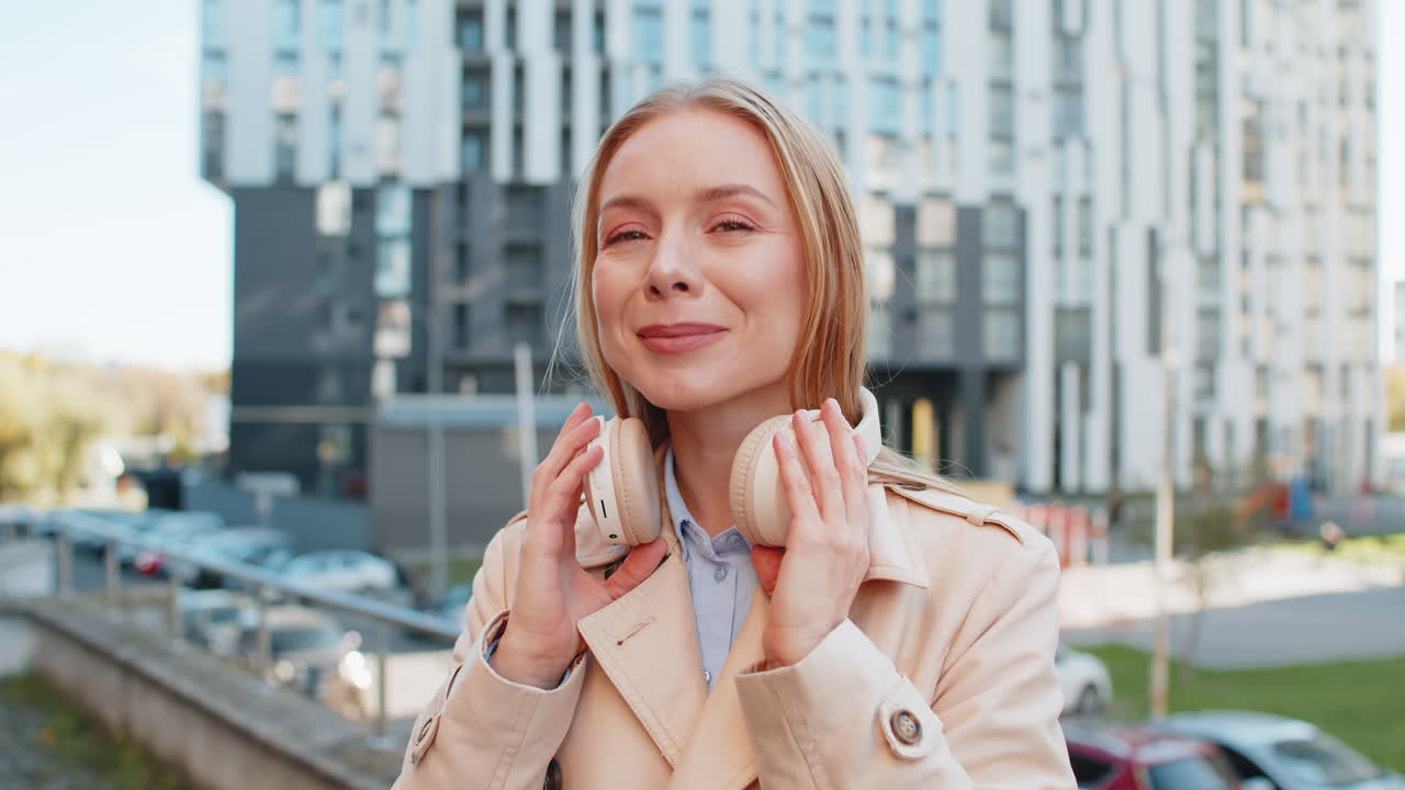 Caucasian mature woman taking off wireless headphones looking at camera and smiling on city street