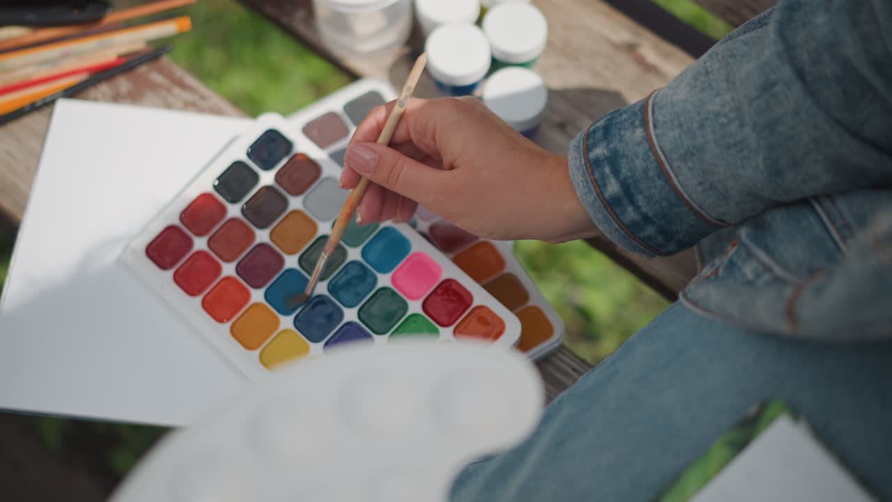 Closeup watercolor palette hand selecting colors on wooden bench with denim jacket, painter dips brush into teal swatch, jars of water and brushes nearby, natural light, intimate creative moment
