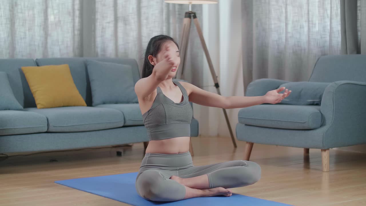 스포츠 옷을 입은 젊은 아시아 운동선수 여성이 요가 매트에 앉아서 집에서 어깨 크로스 팔 스트레칭을 하고 있습니다.