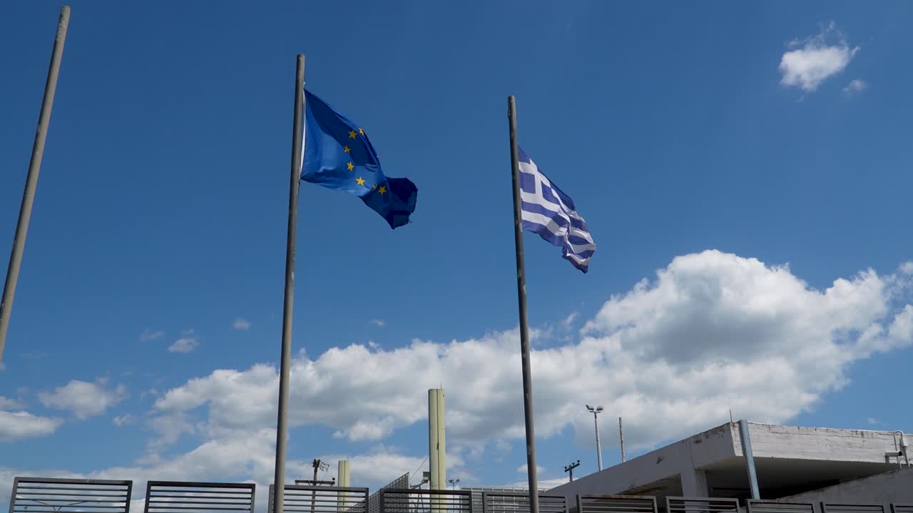 Flags of the European Union and Greece wave on tall poles under a vibrant blue sky with scattered clouds
