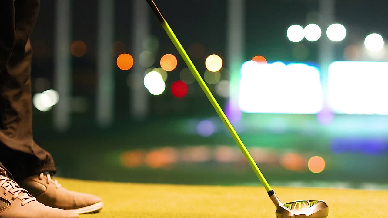 A golfer's shoes and club poised on a lit driving range at night.