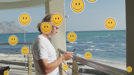 Animation-Von-Social-Media-Emojis-über-Einem-Mann,-Der-Sein-Smartphone-Am-Meer-Nutzt