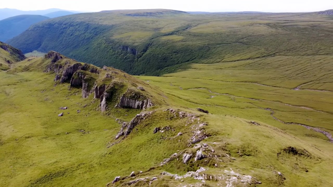 Vista aerea diurna si muove costantemente sopra un terreno verde e aspro, rivelando affioramenti rocciosi lungo una stretta cresta, pareti profonde e in pendenza della valle, deboli canali tortuosi e colline sfocate in lontananza.