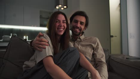 Portrait-of-a-happy-brunette-girl-in-a-beige-T-shirt-together-with-her-brunette-boyfriend-with-stubble-they-sit-on-the-sofa-and-laugh-in-a-modern-apartment-in-the-evening