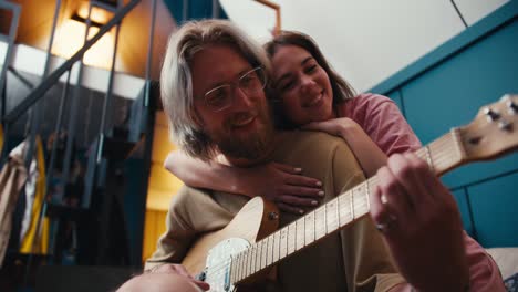 a guy a girl spends time together a blond guy with a beard in glasses plays the electric guitar a brunette girl hugged him and listens