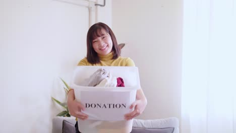 portrait of asian young woman looking at camera holding box with clothes for donation