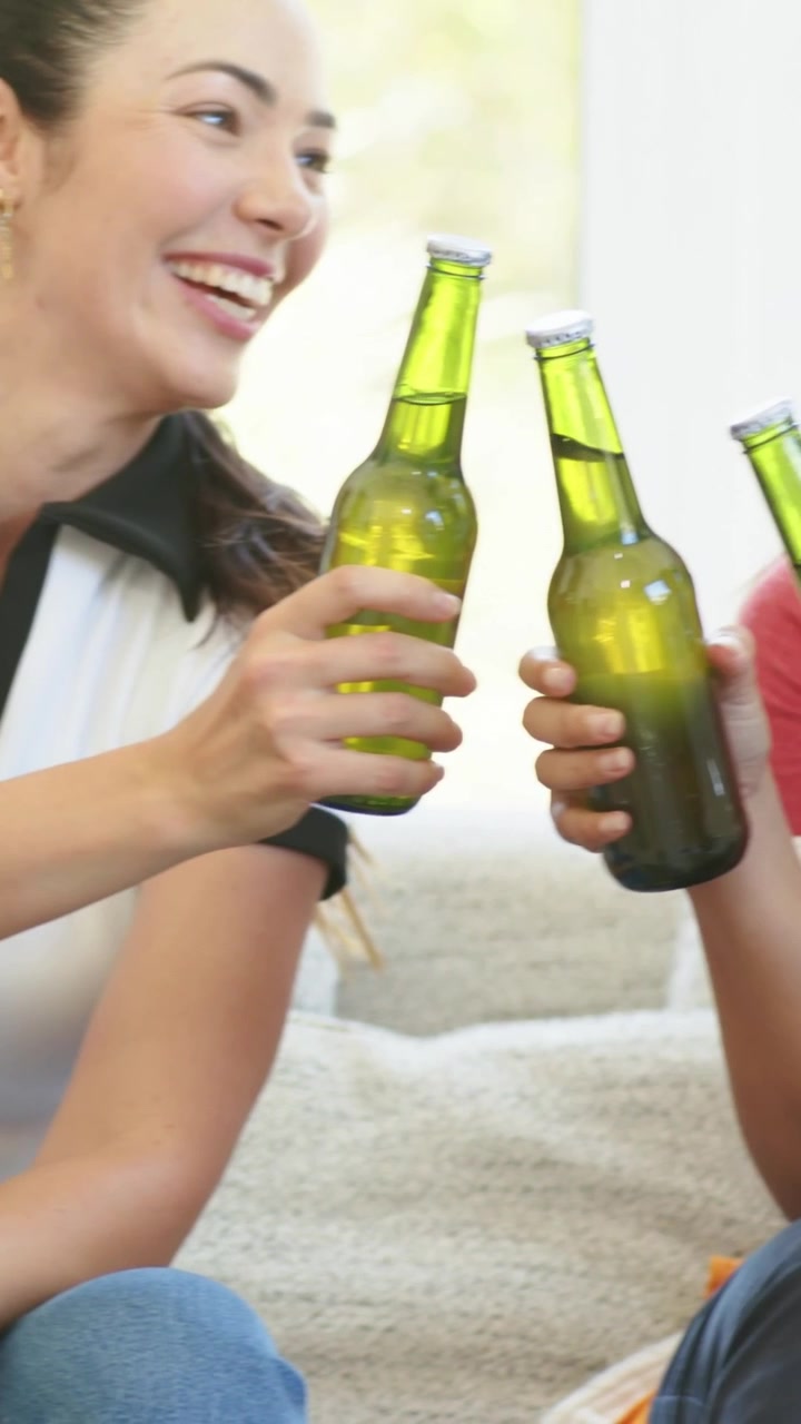 Video vertical: Mujer con polo blanco cogiendo una botella, con las manos alcanzando un paquete de seis en el sofá y brindando.