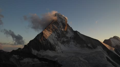 drone shot moving forward towards matterhorn in switzerland with its steep slopes and cliffs filled with snow as the first rays of sun hits in the morning