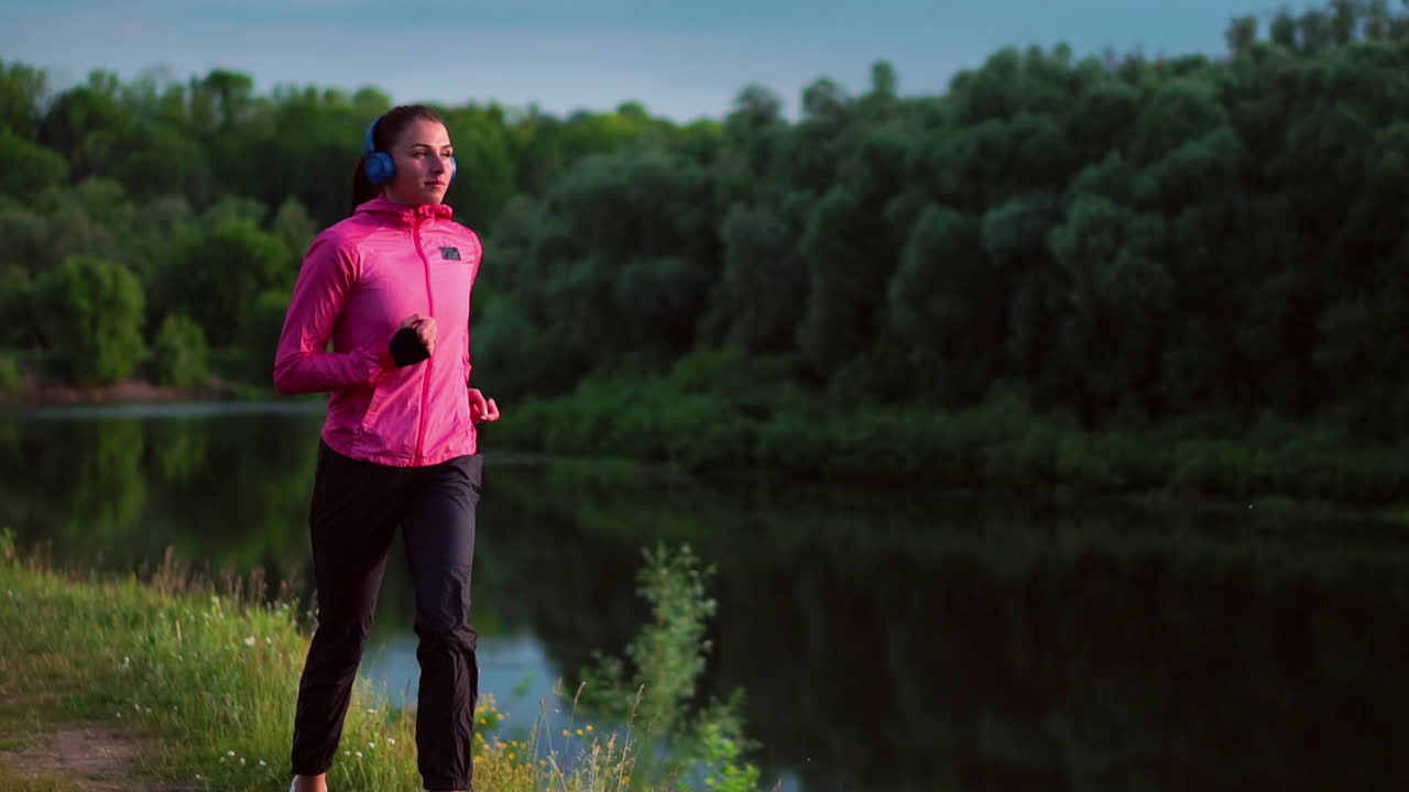 A girl in a pink jacket and black pants runs near the river in headphones preparing for the marathon