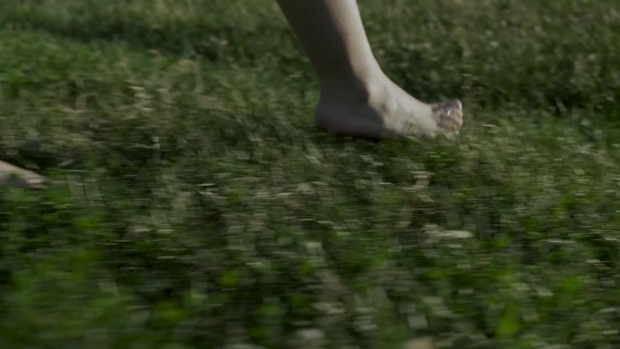 Side shot in slow-motion of a woman walking barefoot on sunny grass and sitting on a blanket