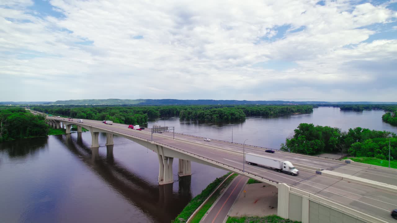 Aerial view of a red semi-truck with a step deck low boy trailer and a dry van trailer crossing the Dresbach Bridge over the Mississippi River at the border of Minnesota and Wisconsin.