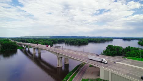 aerial view of a red semi-truck with a step deck low boy trailer and a dry van trailer crossing the dresbach bridge over the mississippi river at the border of minnesota and wisconsin.