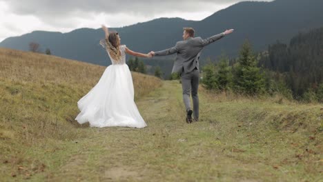 Lovely-young-newlyweds-bride-groom-running-on-mountain-slope,-holding-hands,-wedding-couple-family