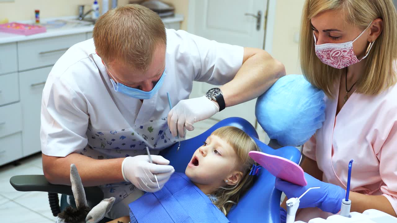 A girl is sitting in a dental chair. The dentist and his assistant treat the teeth of a child in a dental clinic