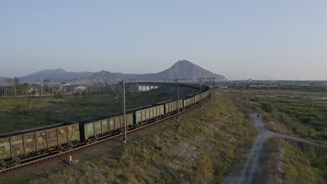 full loaded coal cargo freight train locomotive moving slowly along a high rise railway, in green fields with mountain ridge in the far distance, on the sunset