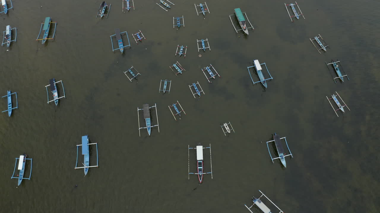 Top-down aerial tilt movement revealing traditional outrigger boats anchored in shallow coastal water in Bali creating a calm maritime composition