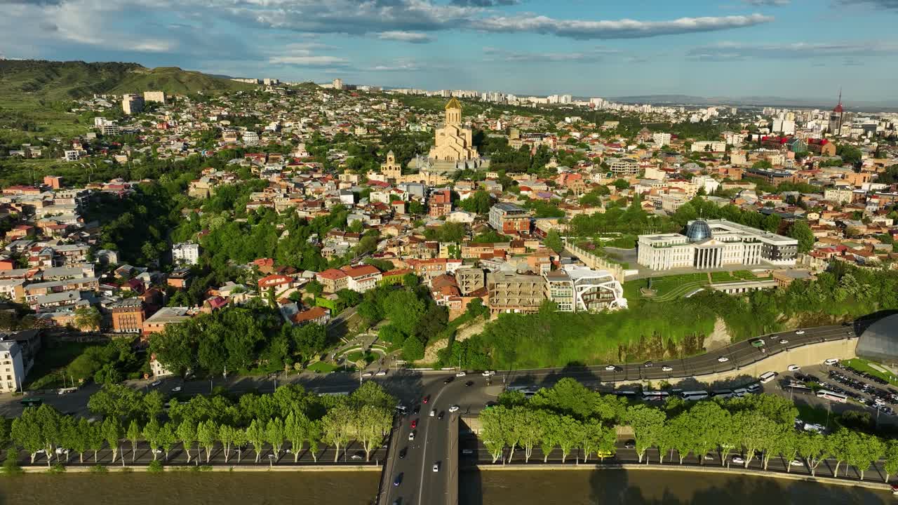 Drone panorama of Old Tbilisi featuring the Holy Trinity Cathedral rising above colorful rooftops, green hills, and the winding Kura River