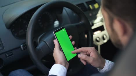 hombre con la mano deslizando la pantalla verde del teléfono inteligente en el volante del coche. joven caucásico borroso navegando por internet en las redes sociales sentado en el asiento del conductor en el vehículo. estilo de vida y tecnologías modernas.