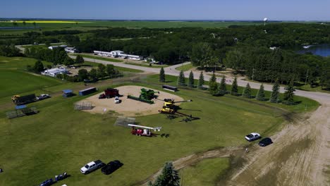 a drone flying towards a rural baseball diamond recreationally area covered in multicolored farm combines tractors and agricultural equipment