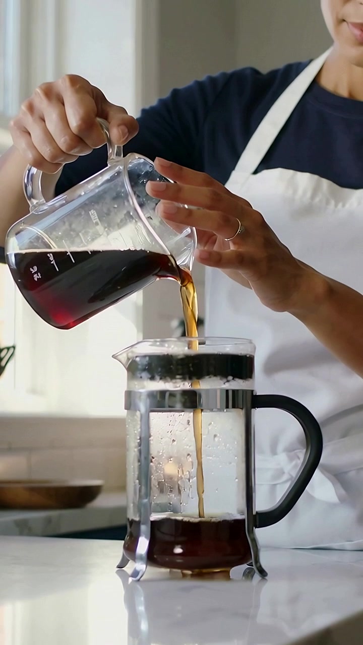 Vídeo vertical: Inclinando jarra de vidro, mulher de avental despejando café na prensa na cozinha para preparar