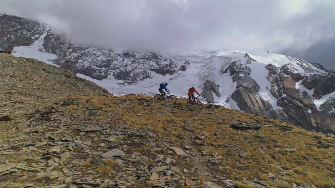 İsviçre'nin Valais kantonunda Saas-Grund ve Saas-Fee Vadisi'nin yukarısında, yüksek bir alp patikasında bisiklet süren iki dağ bisikletçisinin havadan drone çekimi. Destansı manzara, dinamik iniş hareketi ve sinematik macera