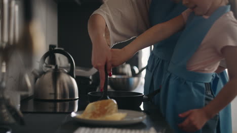 mother is teaching her little daughter to fry pancakes woman and child are standing near stove