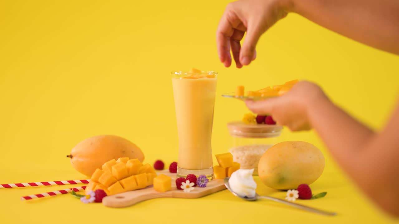 Una mano decora un bicchiere alto di frullato al mango con frutta su uno sfondo giallo brillante da studio