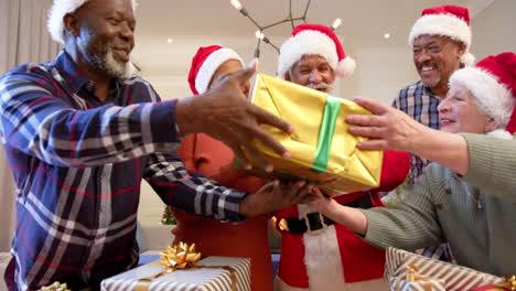 happy diverse senior friends in santa outfit and christmas hats exchanging gift at home, slow motion