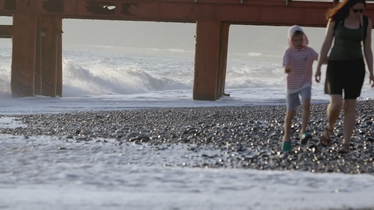 Mother and child walking on a beach with waves and pier