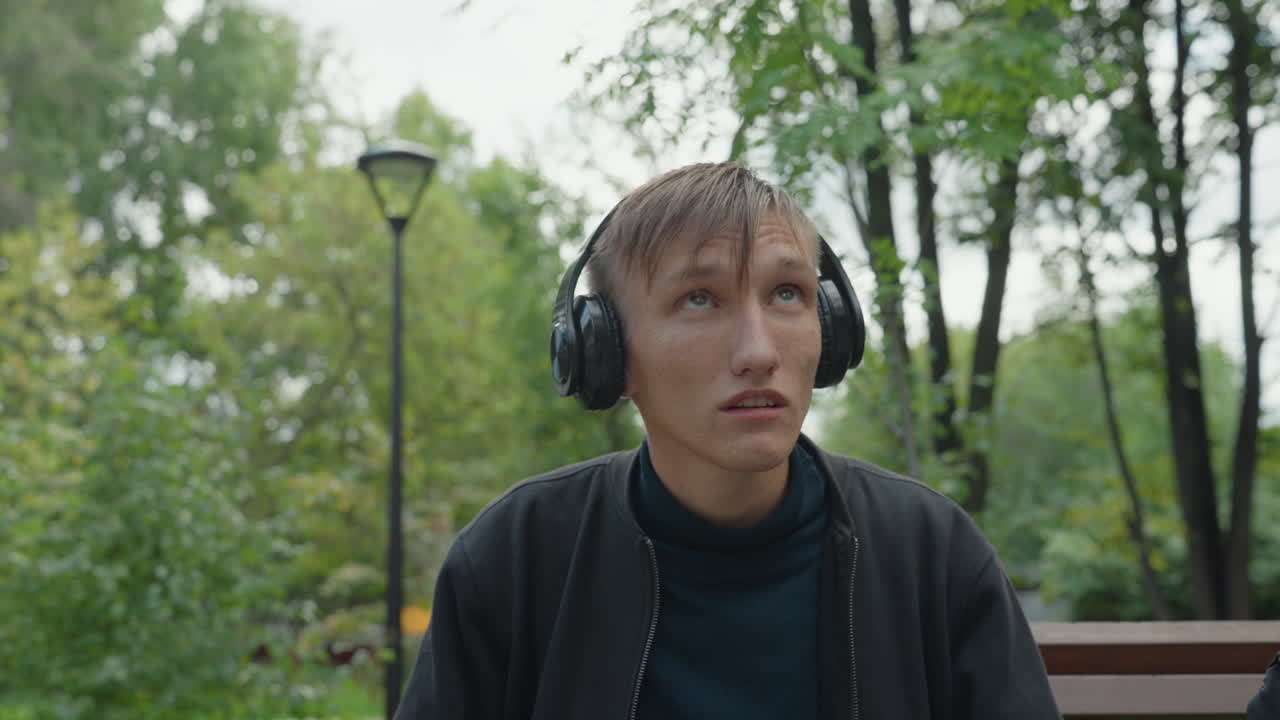 Uomo bianco seduto su una panchina che si sistema le cuffie mentre prova l'audio immersivo in un parco umido, espressione sorpresa con testa inclinata e sguardo rivolto verso il basso, capelli con gocce di pioggia, sfondo con vegetazione verde e lampione