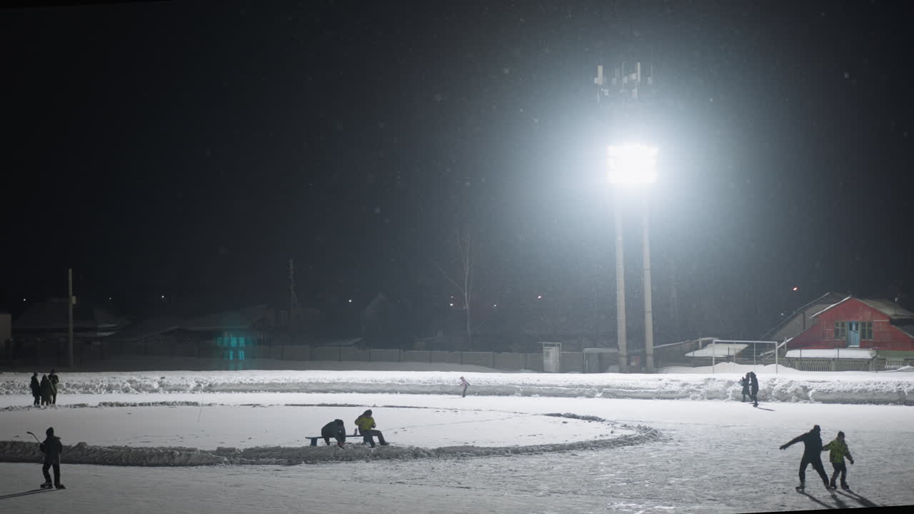 People enjoying winter activity skating on outdoor ice rink at night under intense overhead light, snowflakes falling through cold air, surrounded by snow-covered surroundings