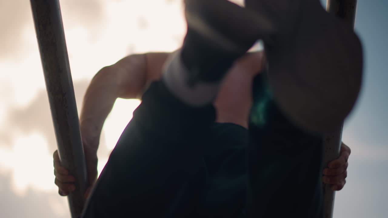 해 질 녘 야외에서 평행봉 운동을 하는 상의를 탈의한 남성, 근육질의 등과 삼두근을 강조한 낮은 앵글의 시네마틱 숏, 쇠봉을 안정적으로 잡고 절제된 동작으로 운동하는 트레이너의 모습, 도시적인 배경