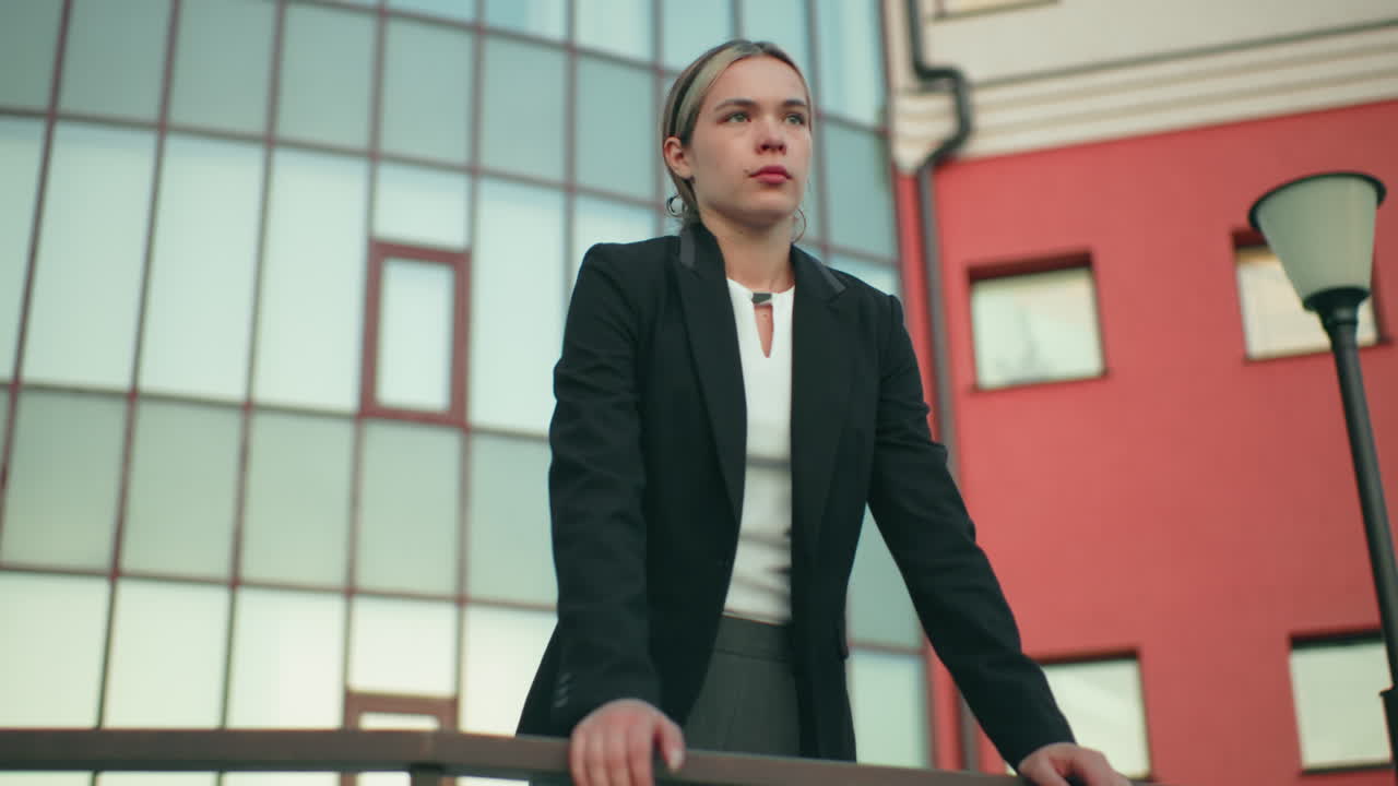 Remote worker in formal outfit leans on iron railing outside residential building, gazing into distance beside lamp post with rooftop light blinking